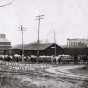 Black and white photograph of horse-drawn wagons hauling bags of seed corn to a railroad station for further transporting, c.1907.