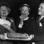 Black and white photograph of the Duchess of Windsor, the former Wallis Simpson, and Philip Pillsbury sample the grand prize winning orange kiss-me-cake cake, 1951.