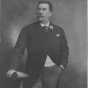 Photograph of Emile Amblard leaning on a table, holding his gloves and wearing a boutonniere. Circa 1910.