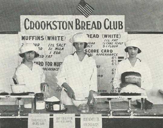 Black and white photograph of three girls in the Crookston Bread Club display baked goods and the recipes used to make them, 1920.