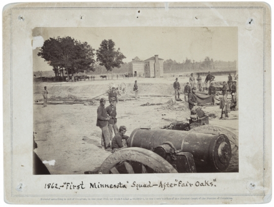 Black and white photograph of men of the First Minnesota Volunteer Infantry in field works after the battlefield of Fair Oaks, Virginia, 1862. Evidence of the newly-attached Company L, the Second Company Minnesota of Sharpshooters, can be seen in the center-right of the photograph: a Sharps breech-loading rifle is hanging over a blanket.